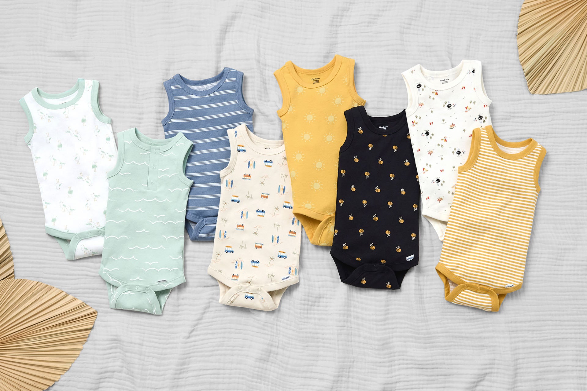 A display of eight sleeveless baby onesies in various colors and patterns arranged on a light-colored fabric background surrounded by decorative leaves.