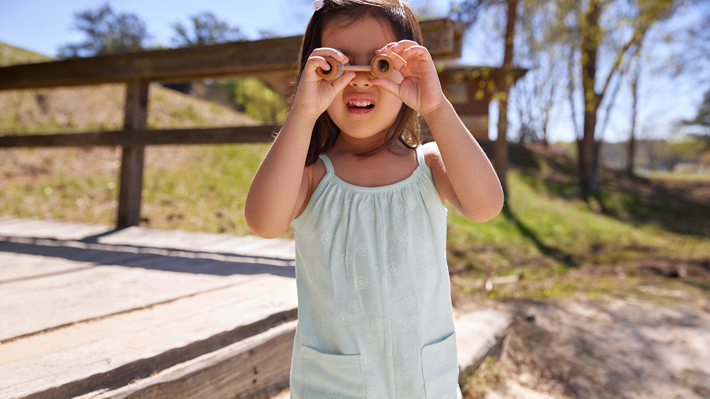 A young girl stands outdoors on a wooden path, holding wooden toy binoculars up to her eyes like glasses. She is wearing a light blue outfit and smiles under a clear, sunny sky with trees in the background.