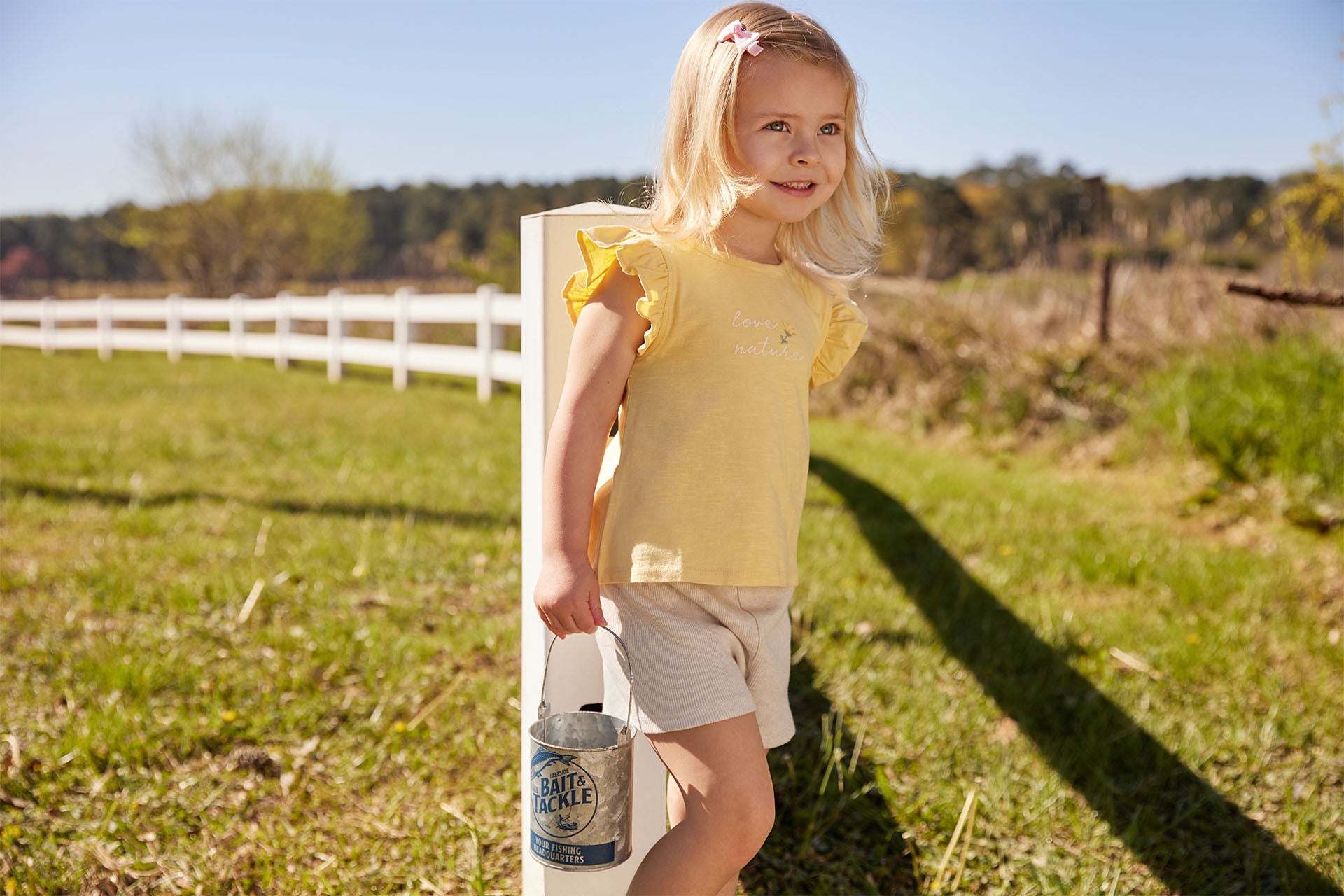 A young girl with blonde hair stands outdoors on a sunny day, holding a small metal bucket. She is wearing a yellow shirt and beige shorts. A white fence and countryside are visible in the background.