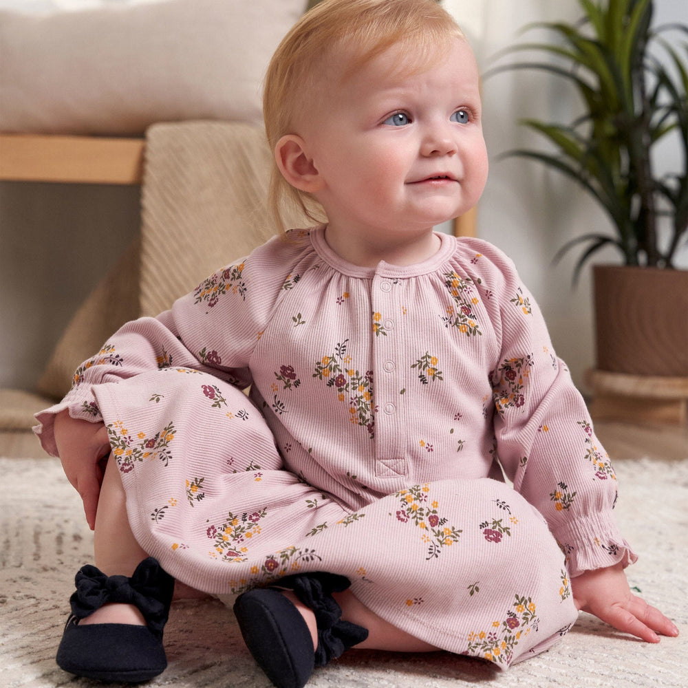 A baby girl in a floral dress sits on a colorful rug, smiling and enjoying her surroundings