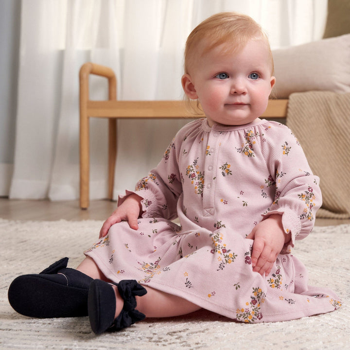 A baby girl in a floral dress sits on a soft rug, surrounded by a cozy and colorful environment