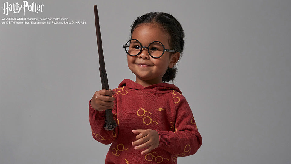 A young girl in round glasses holds a wand, wearing a Harry Potter hoodie adorned with glasses and lightning bolts