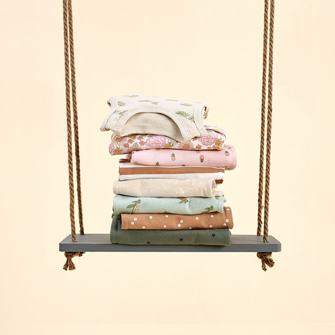 A charming stack of baby bodysuits and clothes in soft pastel colors with delicate patterns, displayed on a rope swing. These must-haves are part of the $5 Frenzy Sale—stock up on essentials while they last!