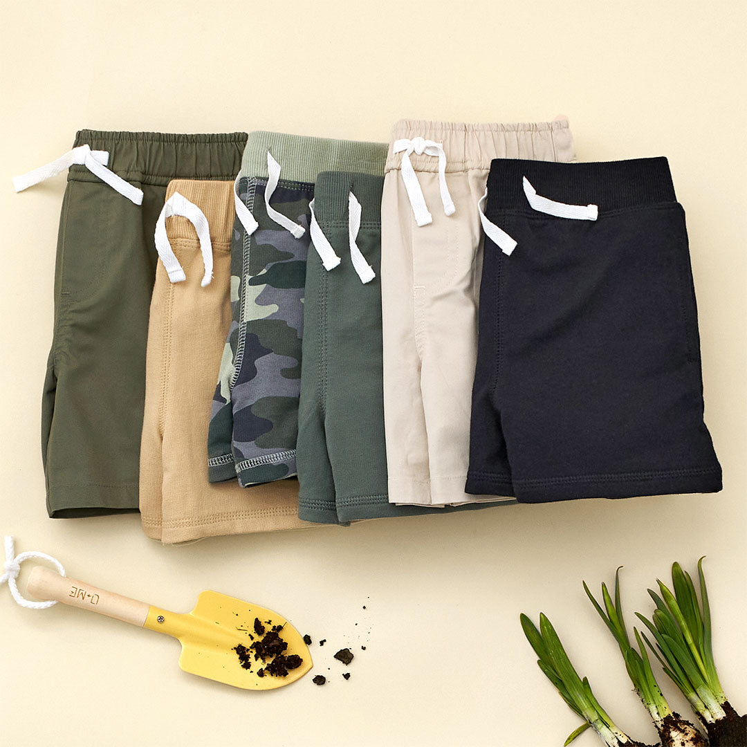 A row of toddler shorts in earthy tones, including camouflage, beige, and black, laid flat on a beige backdrop with gardening props nearby. Mix and match during the $5 Frenzy Sale for unbeatable deals on versatile styles!