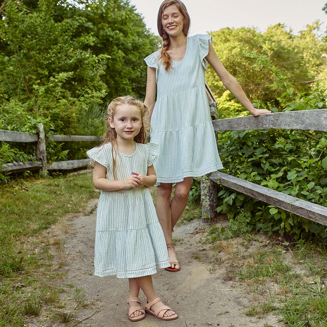 Matching mother-daughter moment with both dressed in soft green gingham dresses, standing along a rustic wooden fence surrounded by greenery. Celebrate family style with deals starting at $14.95!