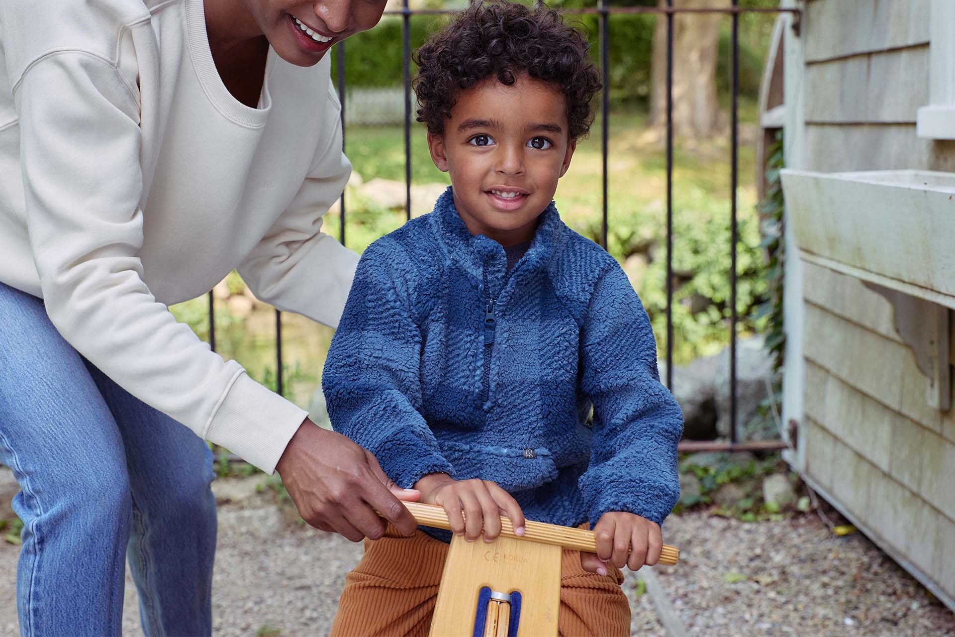 A woman and a child are happily riding a wooden toy, embodying fun and play. Check out our boy toddler clothing collection.