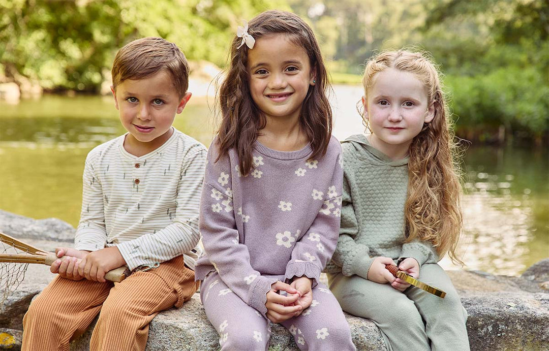 Three children sitting on a rock by a serene lake wearing comfortable, stylish outfits from Modern Moments Brand by Gerber. The boy on the left is holding a net, while the two girls wear matching cozy sets, smiling warmly.