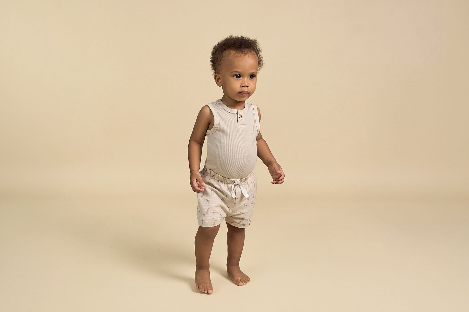 A baby boy wearing Gerber Childrenswear® modern moments™ sleeveless top and matching shorts in neutral tones, standing confidently against a minimalist beige background.