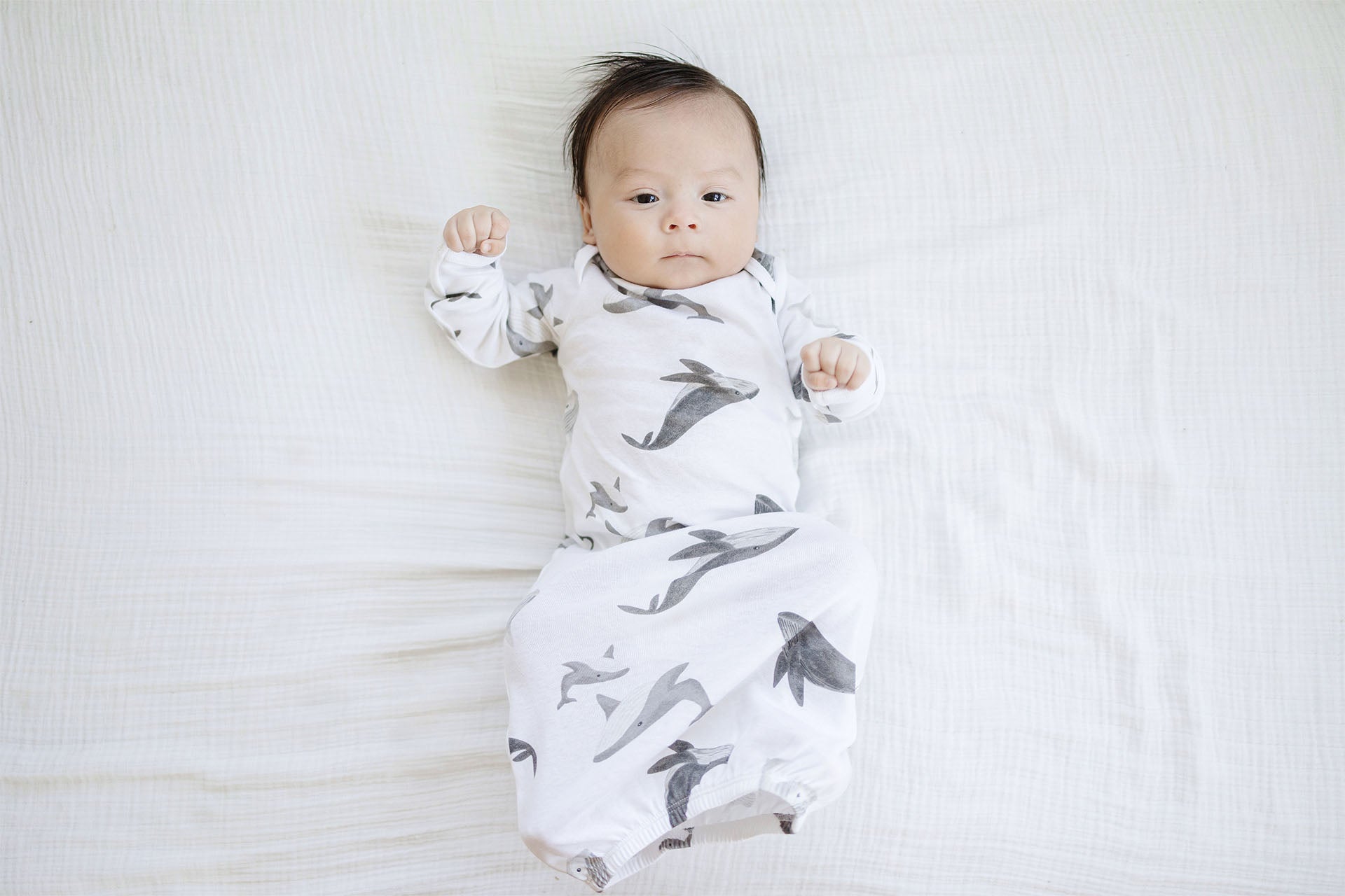 A baby boy lying on a soft white blanket, wearing Gerber Childrenswear® pajamas featuring an adorable gray whale pattern, perfect for cozy springtime naps.”