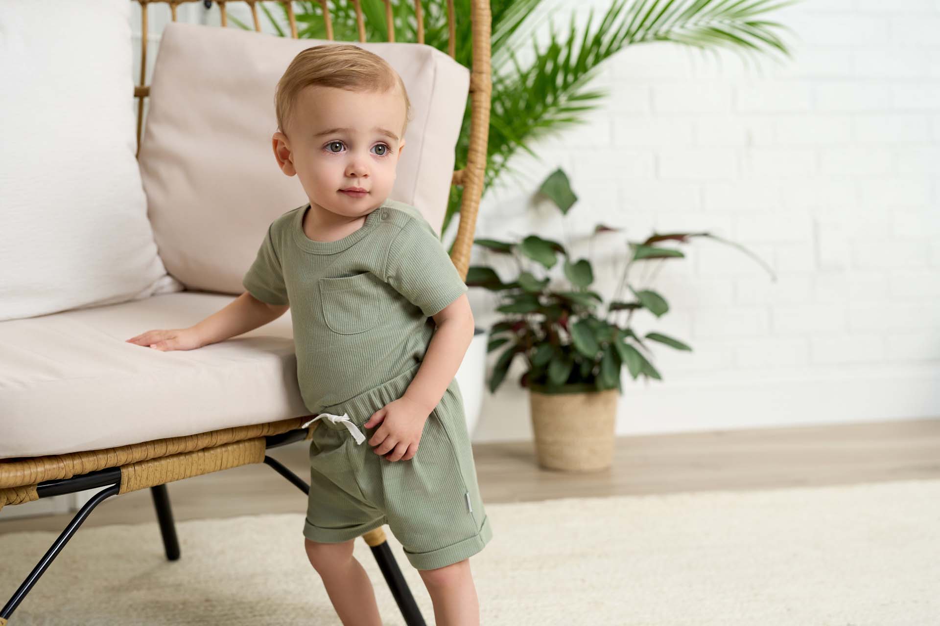 A baby boy wearing a Gerber Childrenswear® olive-green short-sleeve top and matching shorts, leaning on a wicker chair with lush greenery in a bright, airy room.