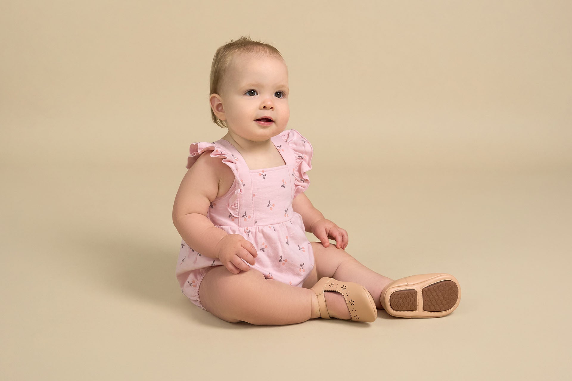 A baby girl sits on a neutral background, wearing Gerber Childrenswear® modern moments™ pink romper with delicate floral prints and ruffled straps, paired with soft, beige baby shoes for a charming spring look.”