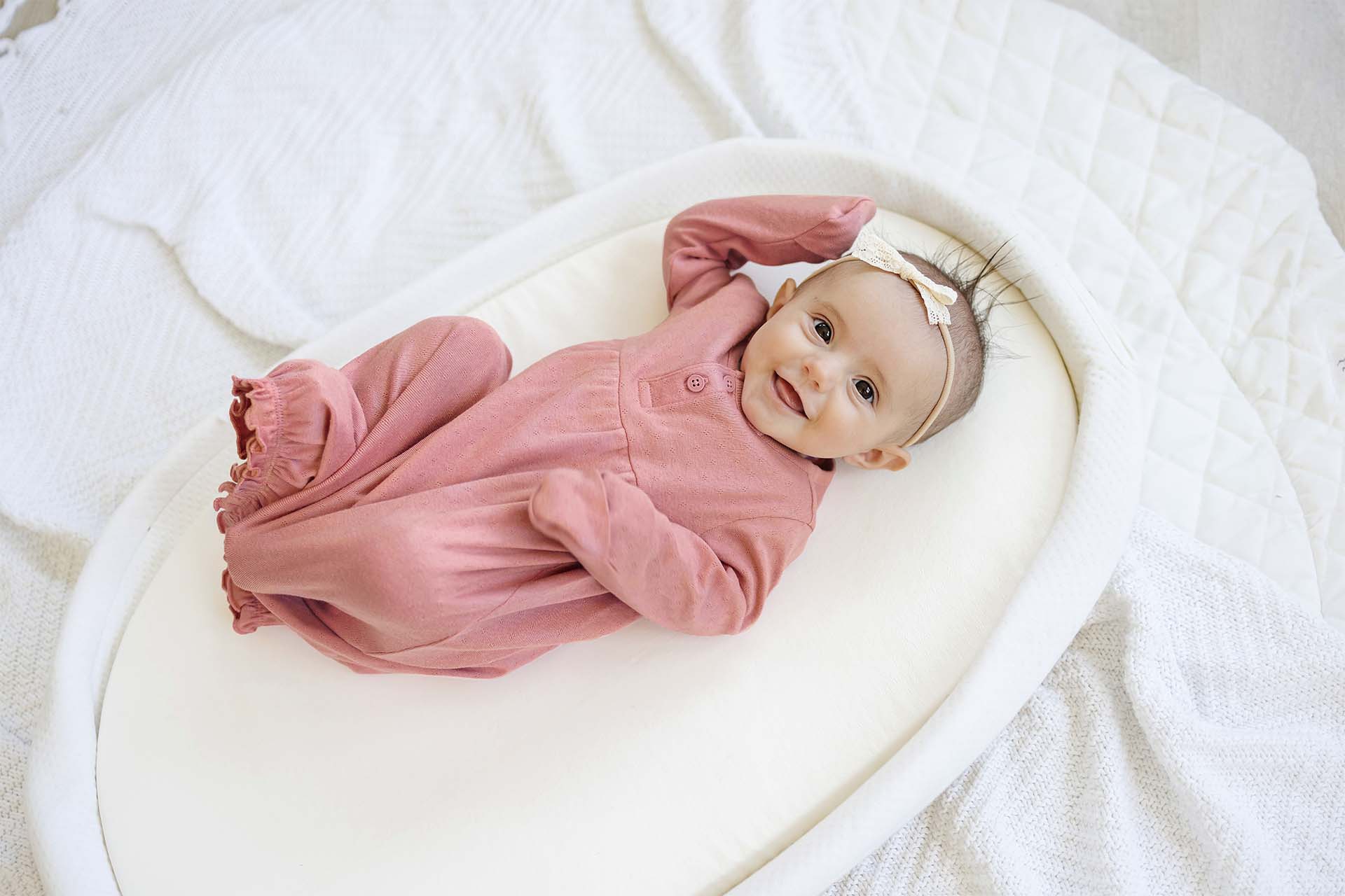 A smiling baby girl wearing Gerber Childrenswear® pink gown style pajamas with ruffle details, lounging on a soft, quilted surface, perfect for cozy springtime naps.