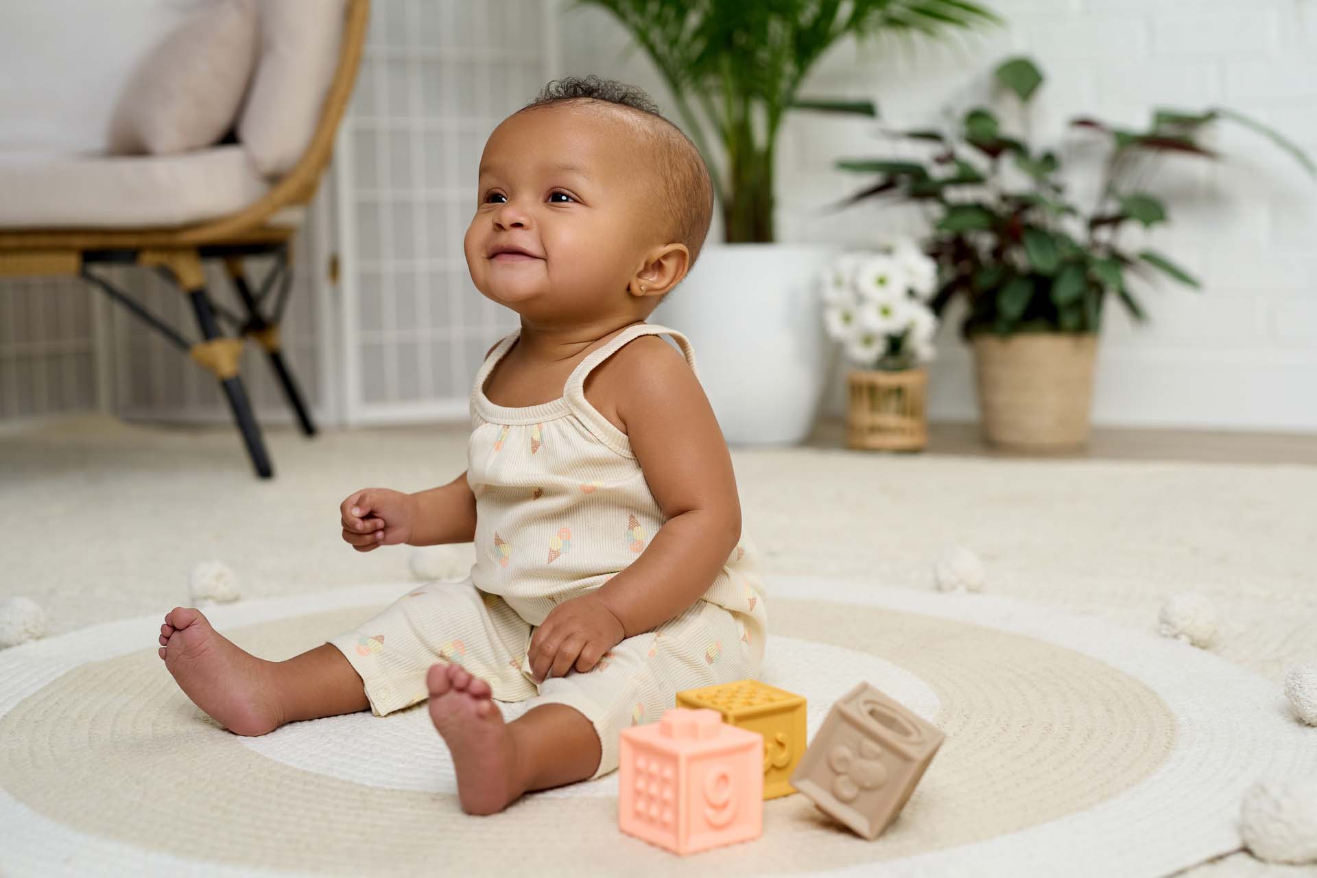 A happy baby wearing Gerber Childrenswear® cream tank romper with subtle ice cream cone prints, sitting on a textured play mat surrounded by soft pastel toys in a bright, airy room.