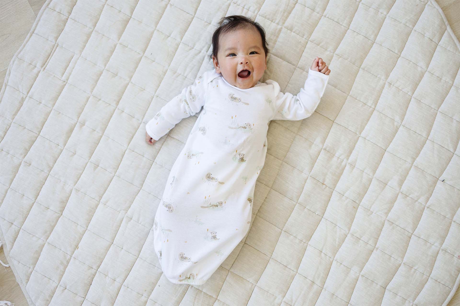 A smiling baby wearing Gerber Childrenswear® white gown style pajamas with an adorable animal print, lying on a soft, quilted blanket for a perfect spring nap.