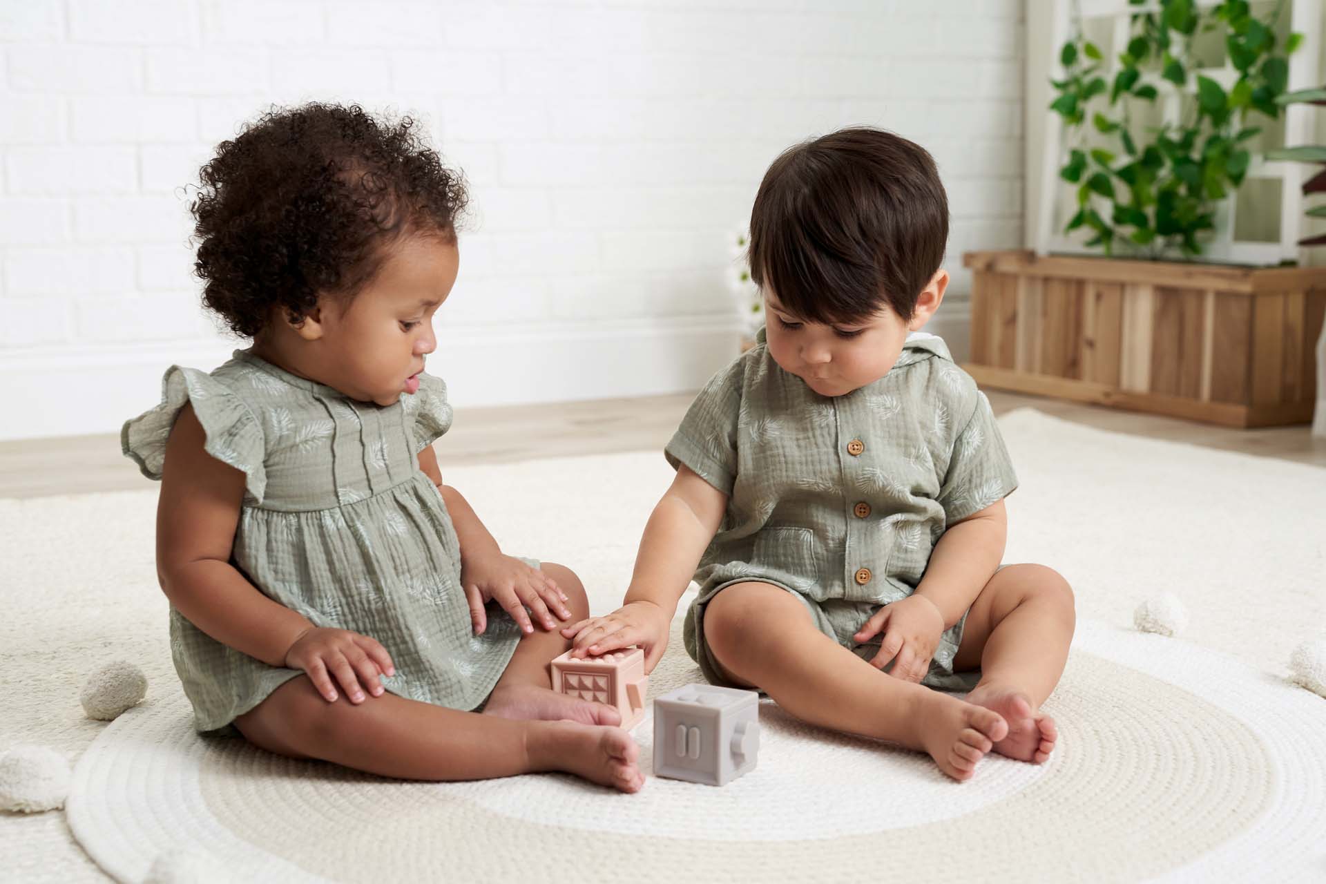 Two babies dressed in Gerber Childrenswear® green muslin outfits sit on a neutral play mat, engaging with pastel blocks in a bright and cozy room.