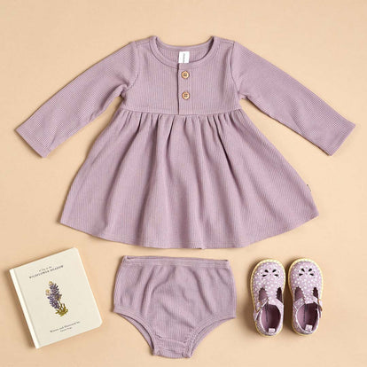  A flat lay of a Gerber Childrenswear® modern moments™ lavender waffle-knit dress with matching bloomers, floral sandals, and a book, perfect for effortless spring style.