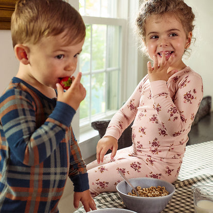  Two toddlers enjoy breakfast in Gerber Childrenswear® modern moments™ pajamas—one in a pink floral print and the other in cozy plaid—adding style to their morning routine.