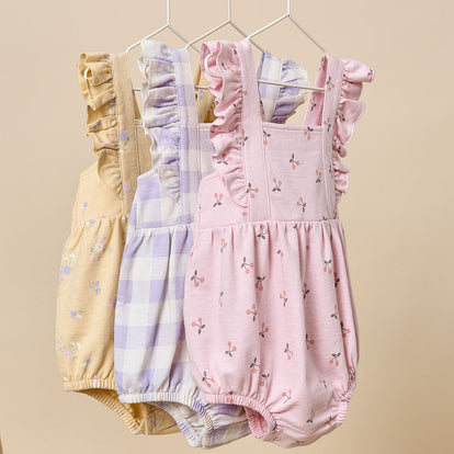  A trio of Gerber Childrenswear® modern moments™ rompers in pastel yellow floral, lavender gingham, and pink cherry prints with ruffle details, perfect for springtime fun.