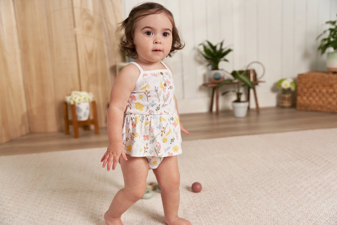 A baby girl wearing a Gerber Childrenswear® floral-print skirted Onesies® Bodysuit with spaghetti straps explores a cozy, plant-filled room, showcasing a playful spring outfit perfect for active little ones.