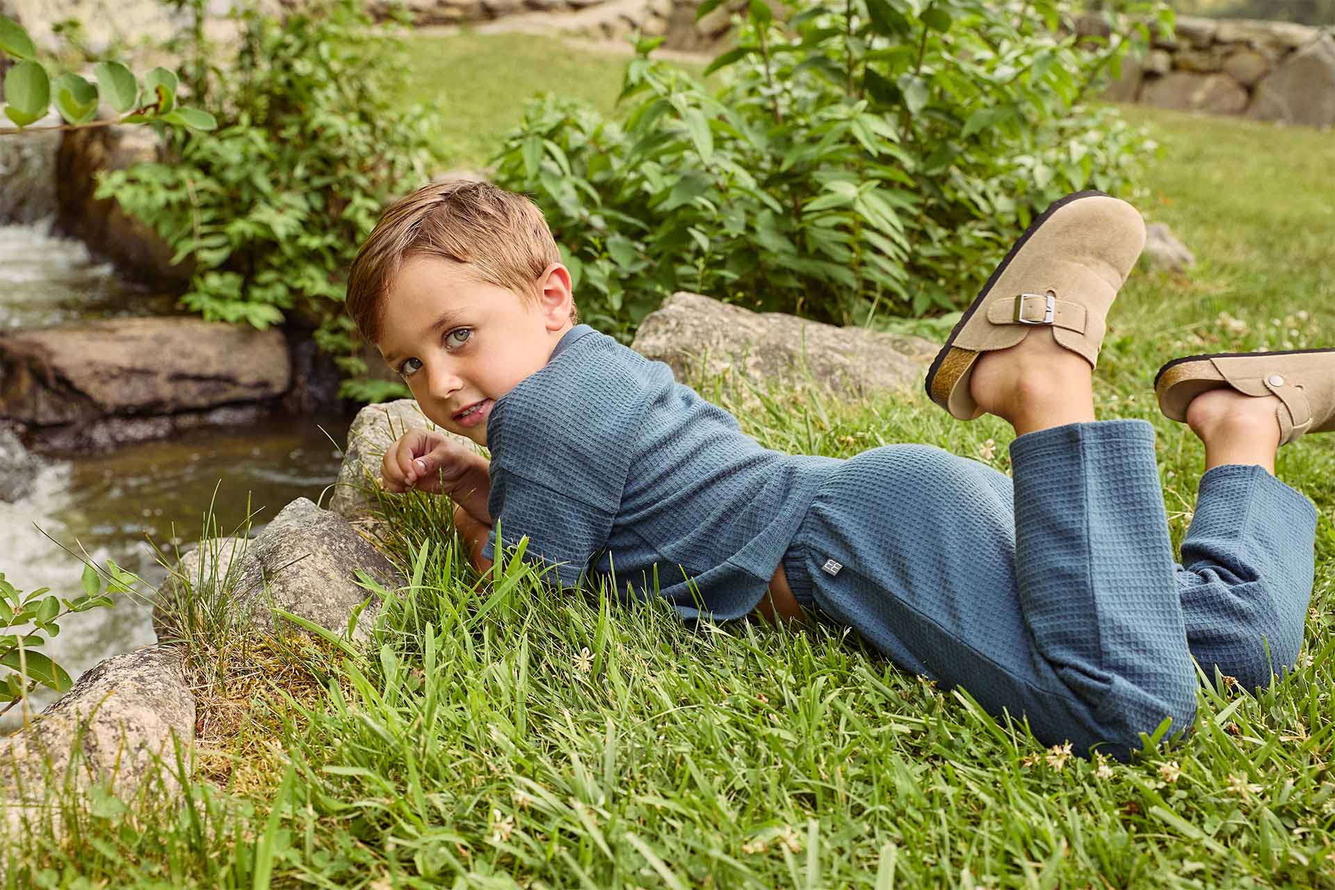 A boy dressed in Gerber Childrenswear® modern moments™ cozy blue outfit relaxes near a scenic stream, enjoying the peaceful outdoors.