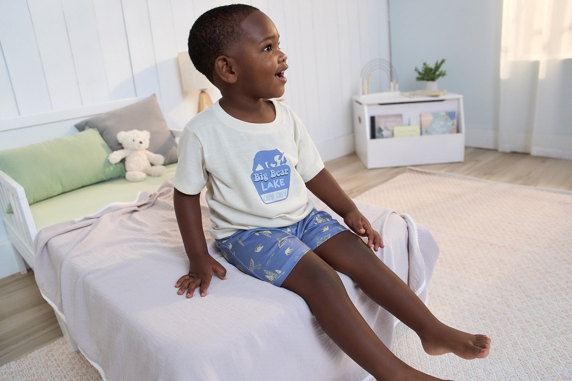 A cheerful toddler boy wearing Gerber Childrenswear® 'Big Bear Lake' pajamas sits on a cozy bed in a softly lit, spring-inspired bedroom.