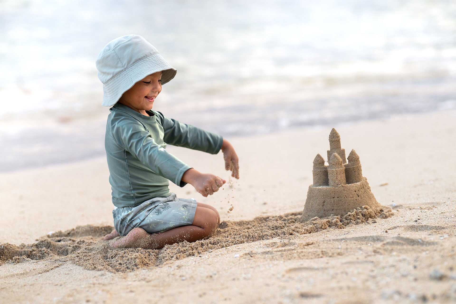 A smiling toddler wearing Gerber Childrenswear® long-sleeve swim top and shorts builds a sandcastle on a sunny beach, perfect for spring adventures.