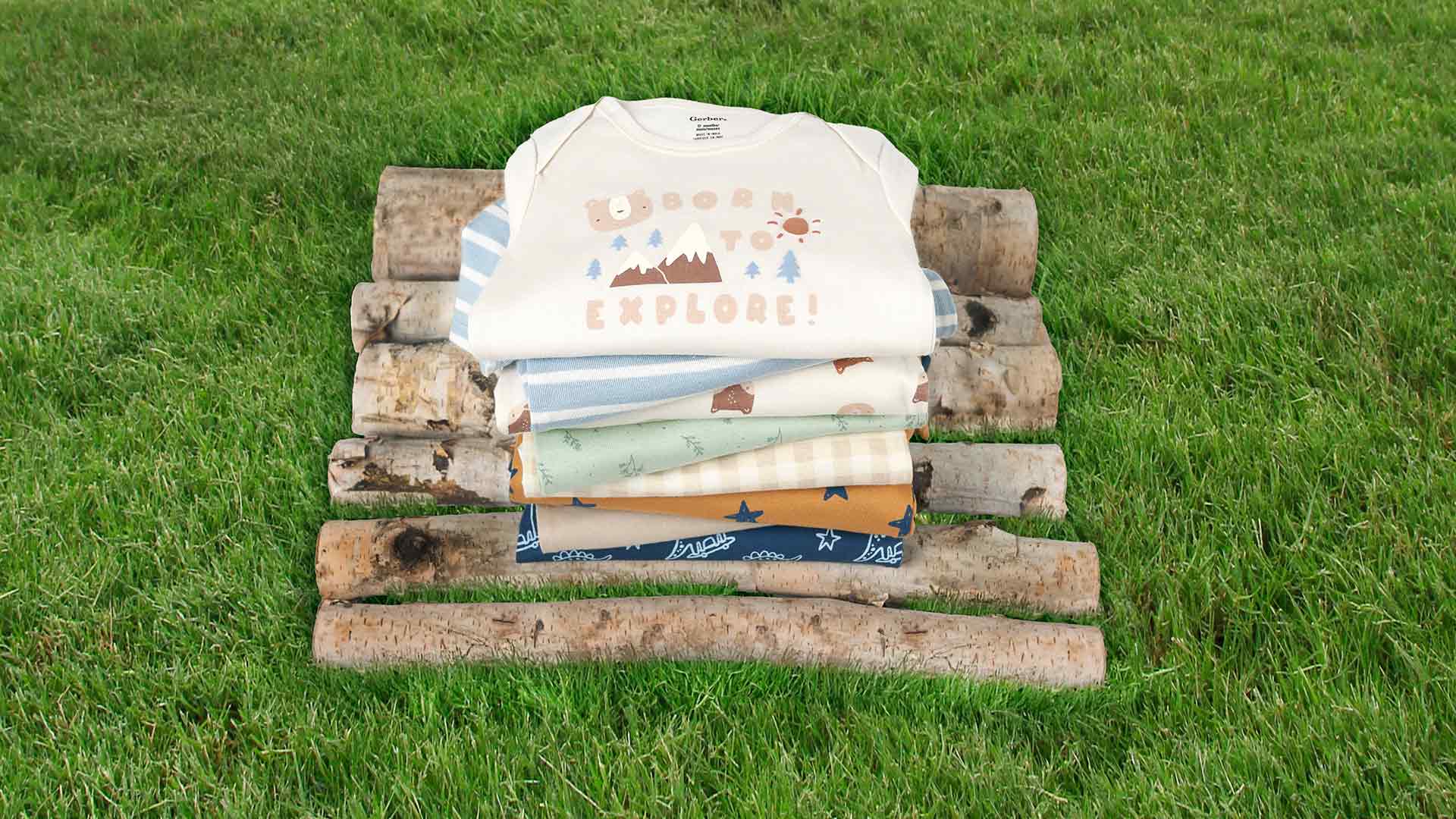 A stack of Gerber Childrenswear® ‘Born to Explore’ themed bodysuits featuring playful outdoor prints, neatly arranged on logs against a grassy background.