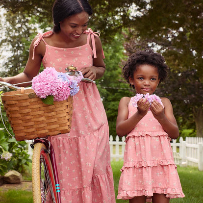  A mother and daughter wearing matching Gerber Childrenswear® pink floral dresses share a moment outdoors, with a bike basket filled with pastel hydrangeas, perfect for springtime outings.