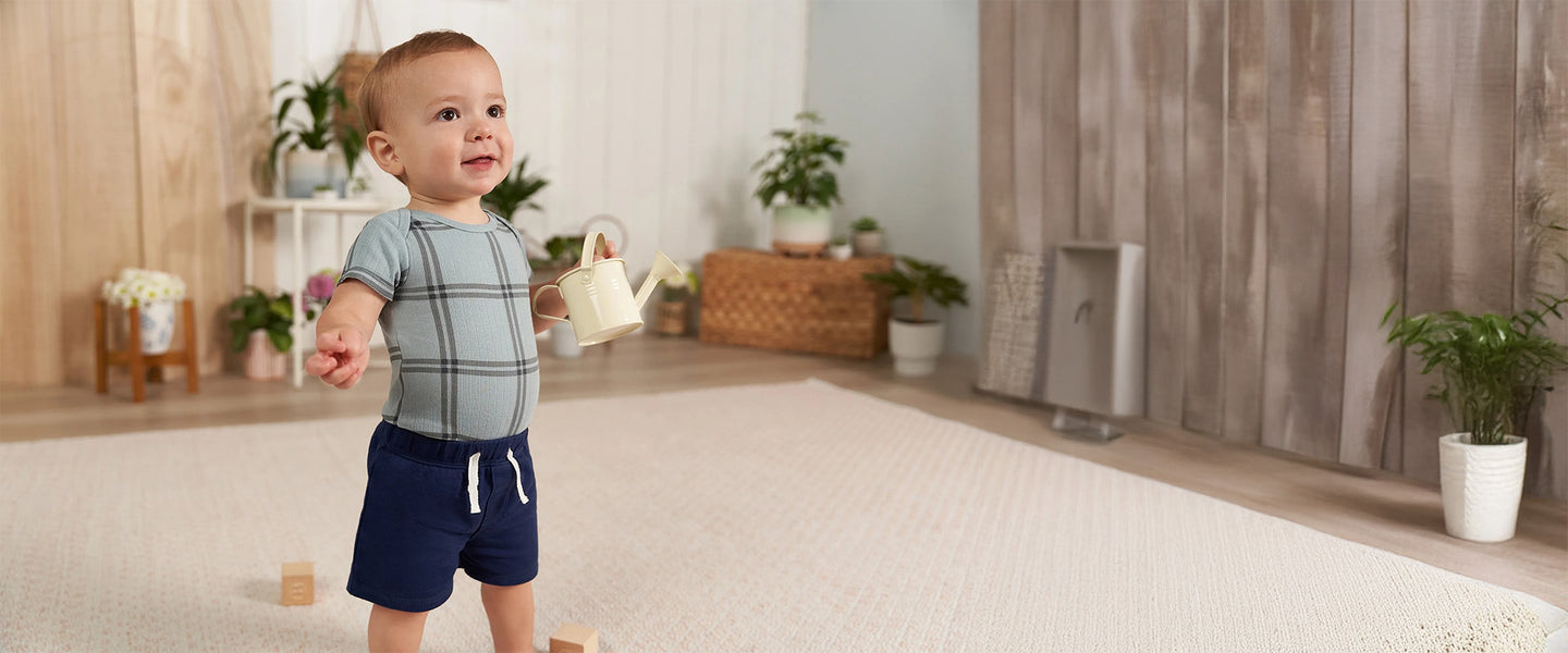 A cheerful baby boy holding a yellow watering can, dressed in a Gerber Childrenswear® plaid bodysuit and navy drawstring shorts, ready for spring playtime.