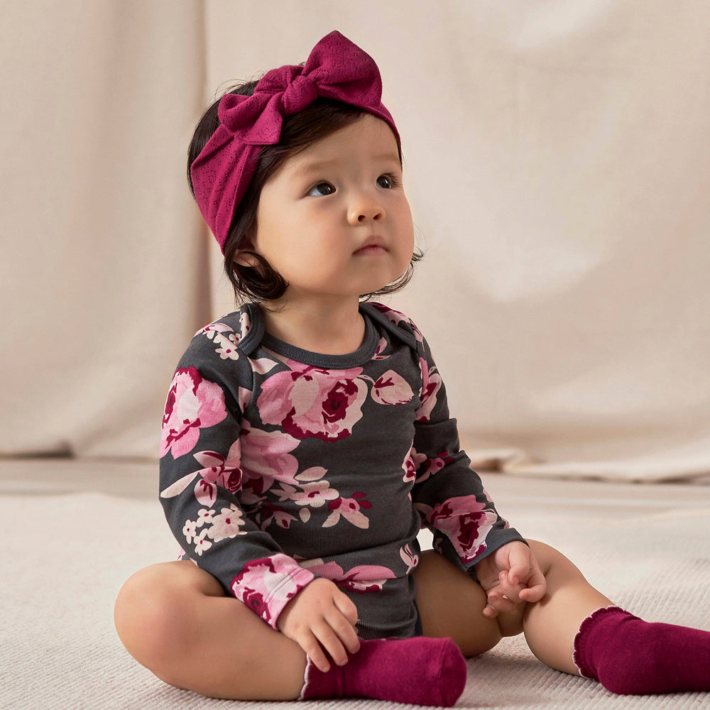 Toddler Girl in a dark gray onesie with a pink floral pattern, matching socks, and a headband with a bow, sits on a light-colored surface against a neutral backdrop of soft fabric.